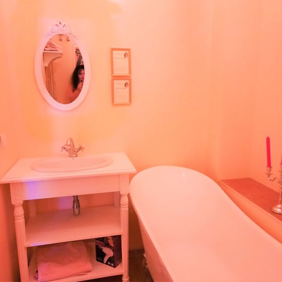 Ein Badezimmer mit einem weißen Waschbecken, Spiegel und Badewanne. Ein rosa Handtuch liegt auf dem unteren Regal des Waschbeckens. Zwei Kerzen befinden sich auf der rechten Seite.