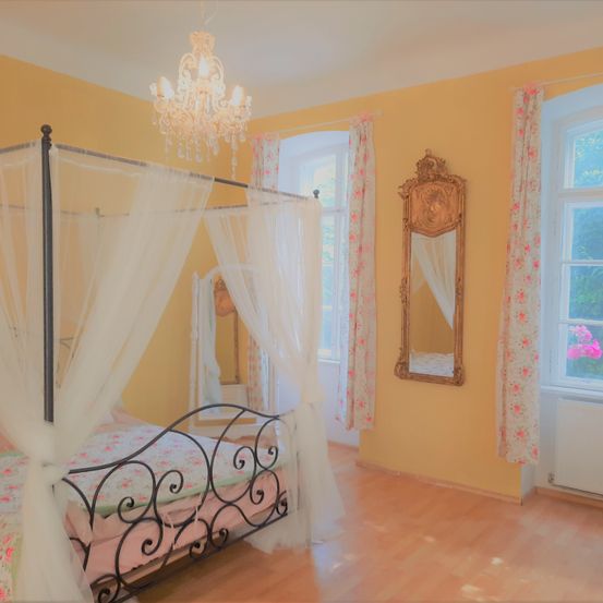 Ein Schlafzimmer mit gelben Wänden, einem Himmelbett mit weißen Vorhängen, einer Kristallleuchte und einem Spiegel mit Blumenvorhängen.