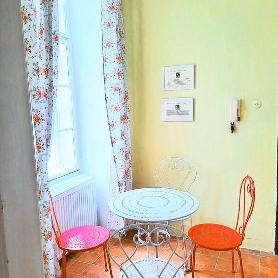 Ein kleiner Essbereich mit einem weißen Tisch und vier Stühlen, zwei davon sind pink und zwei rot. Der Raum hat eine gelbe Wand mit Blumenvorhängen und zwei Bilderrahmen. Ein Telefon ist an der Wand montiert.