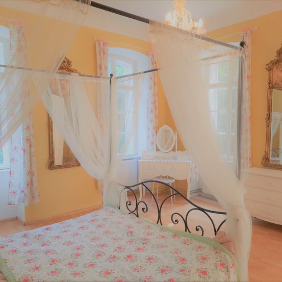 Ein Schlafzimmer mit einem Baldachinbett, gelben Wänden und einem Blumenbettüberwurf. Ein Spiegel und ein Schreibtisch mit Stuhl im Hintergrund.