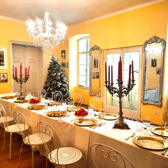 Ein langer Esstisch für einen festlichen Anlass, geschmückt mit goldenen Tellern, roten Kerzen und einem Weihnachtsbaum. Der Raum hat gelbe Wände, eine Kristallleuchte und prunkvolle Spiegel.
