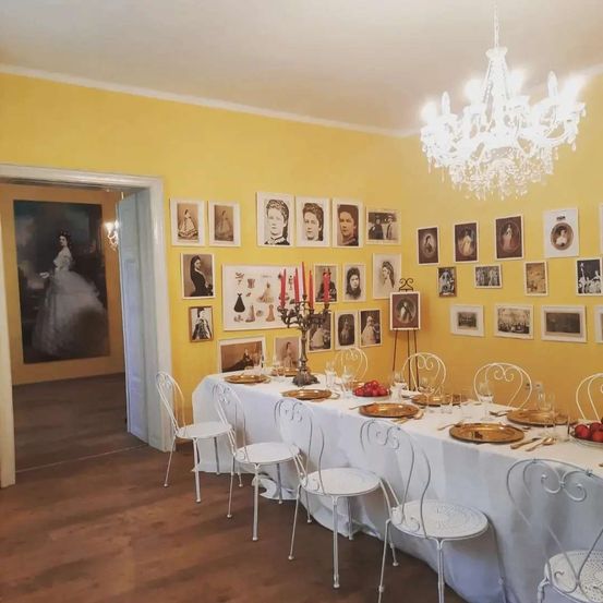 Ein Esszimmer mit einem langen Tisch für eine formelle Veranstaltung, geschmückt mit weißen Stühlen, einer Kristallleuchte und einer gelben Wand, die mit gerahmten Porträts gesäumt ist.