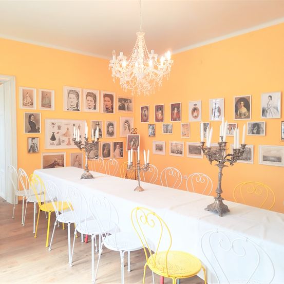 Ein Esszimmer mit einem langen Tisch, der mit Weiß bedeckt ist, umgeben von weißen und gelben Stühlen. Die Wände sind orange gestrichen und mit gerahmten Porträts geschmückt. Zwei Kandelaber mit brennenden Kerzen stehen auf dem Tisch.