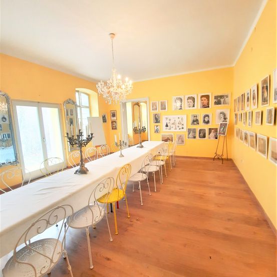 Ein leerer Raum mit gelben Wänden, einem langen Tisch, der mit weißem Tuch bedeckt ist, und verschiedenen gerahmten Bildern an der Wand. Es gibt einen Kronleuchter, einen Kerzenhalter und einen Spiegel.