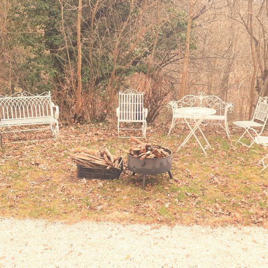 Ein Garten mit einem Feuerkorb, einer Bank und einem Tisch mit Stühlen. Der Bereich ist von Bäumen umgeben.