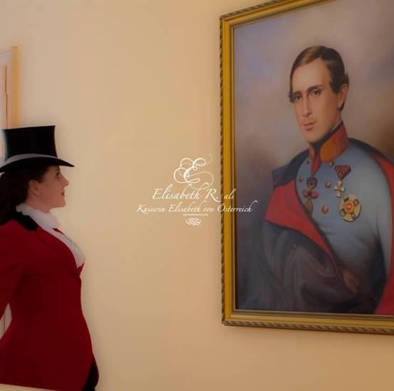 Eine Frau in einem roten Kostüm mit einem schwarzen Hut steht vor einem Porträt eines Mannes in Uniform.