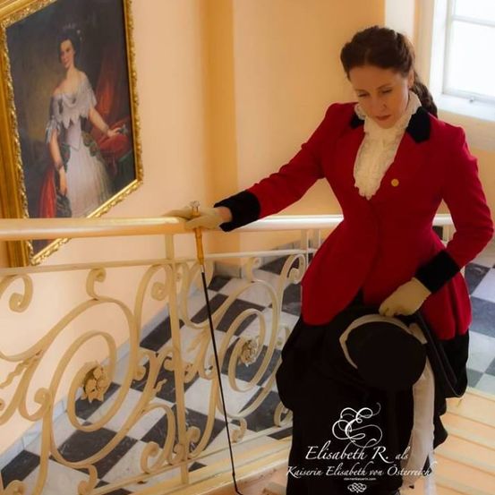 Eine Frau in einem roten Mantel und schwarzem Rock steht auf einer Treppe und hält eine Seil. Ein Porträt einer Frau in einem weißen Kleid hängt an der Wand hinter ihr.