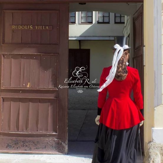 Eine Frau in einem roten und schwarzen Kostüm mit einem weißen Hut steht vor dem Eingang der Rudolfs Villa.