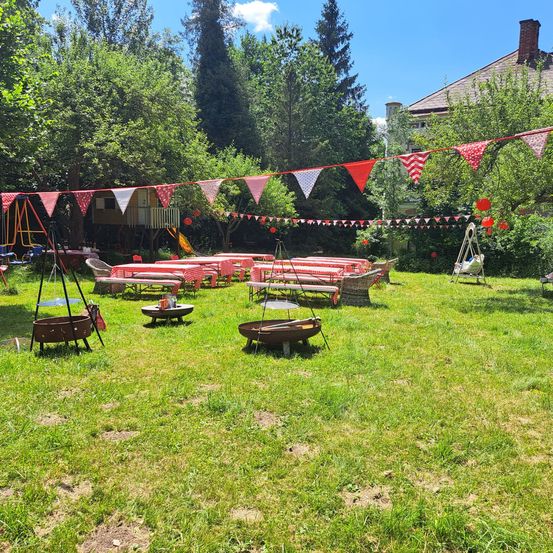 Ein gut organisierter Außenbereich mit in Reihen angeordneten Tischen und Stühlen, dekoriert mit rot-weißer Beflaggung. Es gibt zwei Feuerstellen, eine Schaukel und ein Baumhaus im Hintergrund.