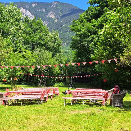 Ein Picknick-Setup auf einer Rasenfläche mit Tischen und Stühlen, rot-weiß karierten Tischdecken und Wimpelketten. Bäume und Berge im Hintergrund.