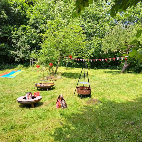 Ein Hinterhof-Setup mit zwei Feuerstellen, einem Grill und bunter Bespannung zwischen Bäumen. Ein bunter Teppich ist auf dem Gras ausgebreitet.