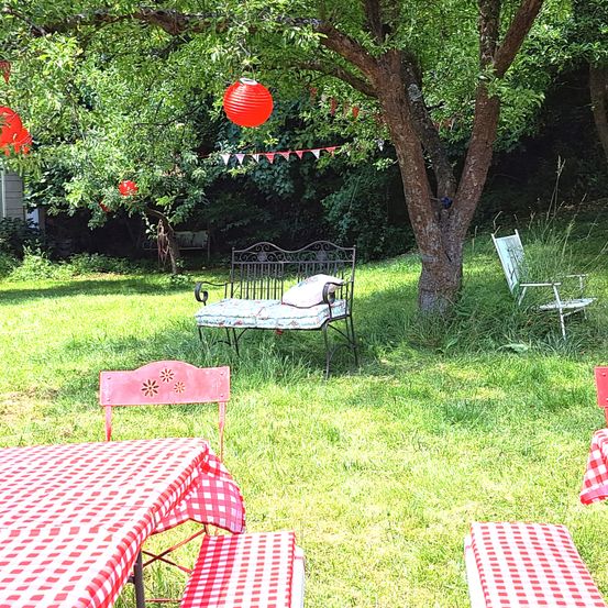 Ein Gartenbereich mit rot-weiß kariertem Tischtuch, einer Bank und einem Baum mit hängenden Laternen.