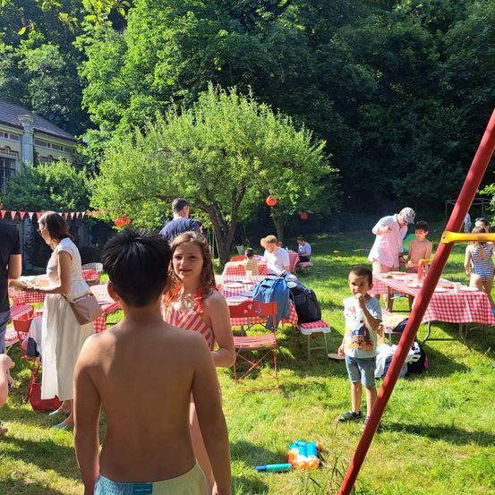 Eine Gruppe von Menschen, einschließlich Kindern, versammelt sich in einem Park für ein Picknick. Tische sind mit rot-weiß karierten Tischdecken gedeckt. Eine Schaukel und ein Baum sind im Hintergrund.