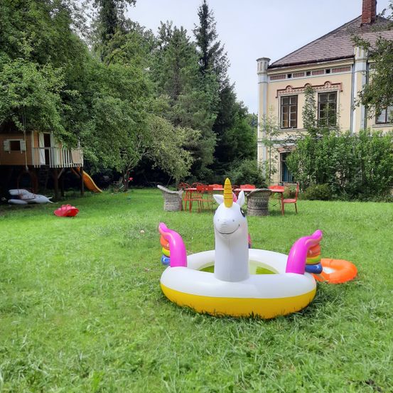 Ein bunter aufblasbarer Einhorn-Schwimmring steht auf einem Rasen vor einem Haus mit Baumhaus und Rutsche.