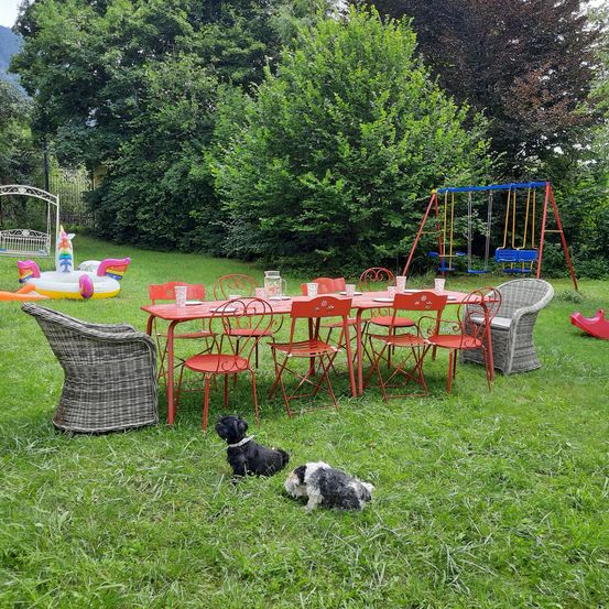 Ein Garten mit einem roten Tisch und Stühlen, einem Schaukelset und einem bunten aufblasbaren Schwimmtier. Zwei Hunde sitzen auf dem Gras.