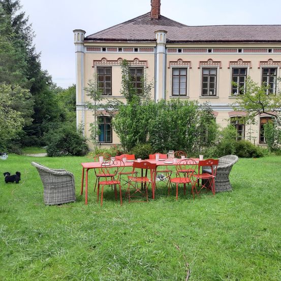 Ein großes, verziertes Gebäude mit mehreren Fenstern steht hinter einem ordentlich arrangierten Esstisch im Freien auf einem grünen Rasen. Ein kleiner schwarzer Hund ist in der Nähe.