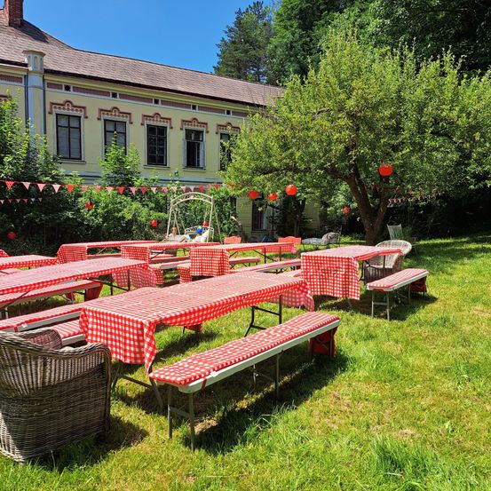 Ein Gartenbereich mit Tischen und Bänken, die mit roten und weißen karierten Tischdecken bedeckt sind, umgeben von Bäumen und einem Gebäude mit roten Laternen, die an seinen Ästen hängen.