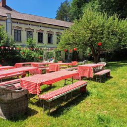 Ein Gartenbereich mit Tischen und Bänken, die mit roten und weißen karierten Tischdecken bedeckt sind, umgeben von Bäumen und einem Gebäude mit roten Laternen, die an seinen Ästen hängen.