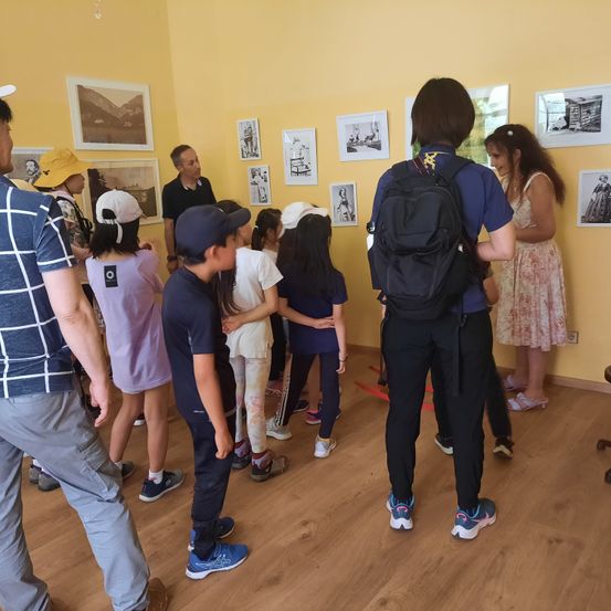 Eine Gruppe von Menschen, einschließlich Kindern, versammelt sich in einem Raum mit gelben Wänden und betrachtet gerahmte Bilder an der Wand. Ein Mann in einem schwarzen Shirt spricht zu ihnen.