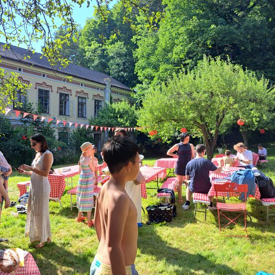 Eine Gruppe von Menschen versammelt sich in einem Garten mit aufgestellten Tischen und Stühlen für ein Picknick. Einige sitzen, andere stehen, und es gibt Dekorationen über den Tischen.