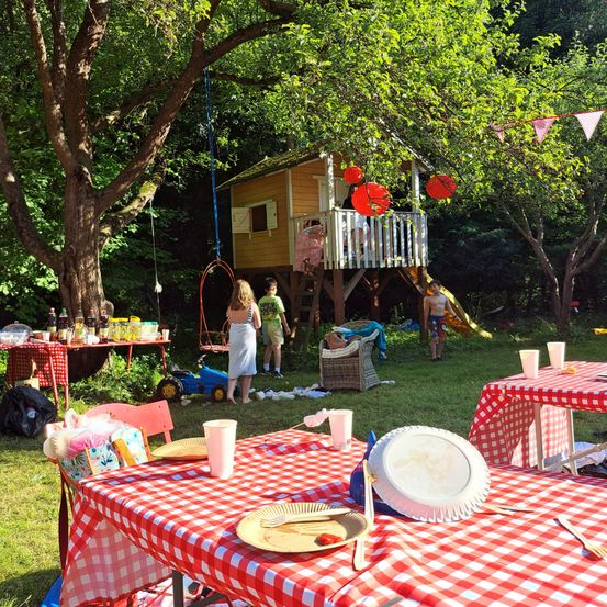 Ein Gartenfest mit einem Baumhaus, Tischen mit rot-weiß karierten Tischdecken und Kindern, die im Hintergrund spielen.