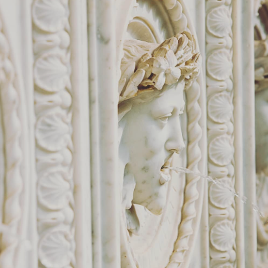Ein Nahaufnahme einer Marmorwand mit einer Brunnenfigur, die Wasser abgibt, umgeben von kunstvollen Ornamenten.