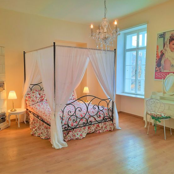 Ein Schlafzimmer mit einem Himmelbett, Blumenbettwäsche und einer Kristallleuchte über dem Bett. Zwei Lampen flankieren das Bett, und eine Kommode mit einem Stuhl und einem Spiegel steht rechts.