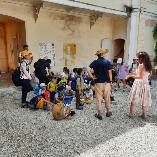 Eine Gruppe von Kindern und Erwachsenen versammelt sich vor einem Gebäude, einige sitzend und andere stehend, mit Rucksäcken und Hüten sichtbar.