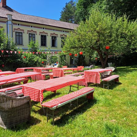 Eine Reihe von Picknicktischen mit rot-weiß karierten Tischdecken in einem Grasbereich mit einem großen Haus im Hintergrund.