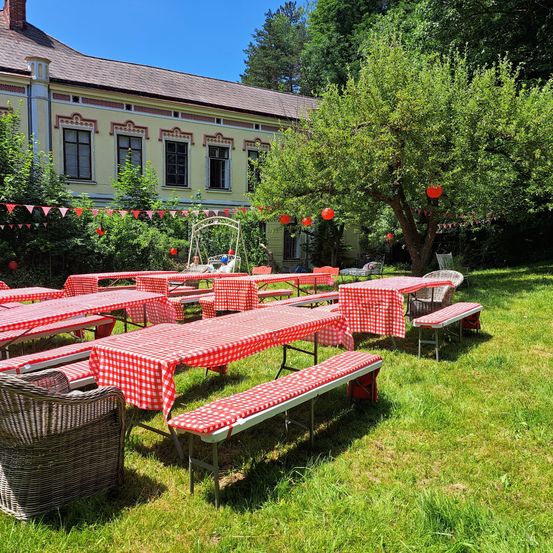Eine Reihe von Picknicktischen mit rot-weiß karierten Tischdecken in einem Grasbereich mit einem großen Haus im Hintergrund.