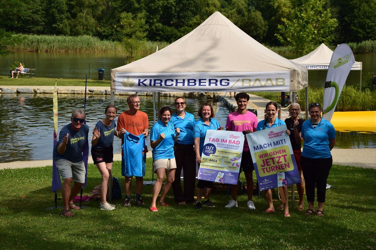 Eine Gruppe von Menschen in passenden blauen Shirts posiert vor einem Zelt mit einem See im Hintergrund. Ein Mann hält ein blaues Shirt. Zwei Frauen halten Schilder, die 'Tag im Bad' und 'Mach mit!' sagen.