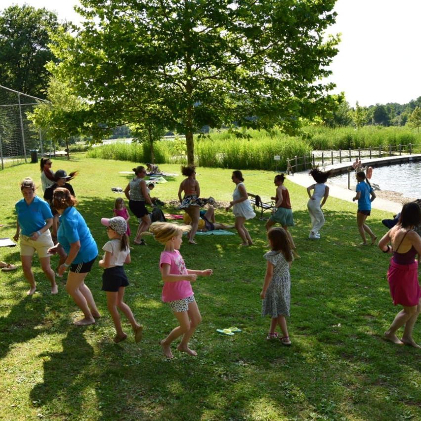 Eine Gruppe von Menschen, einschließlich Kindern, spielt auf einem Rasenfeld in der Nähe eines Sees. Einige sind barfuß, während andere Sandalen tragen. Ein Baum spendet Schatten.