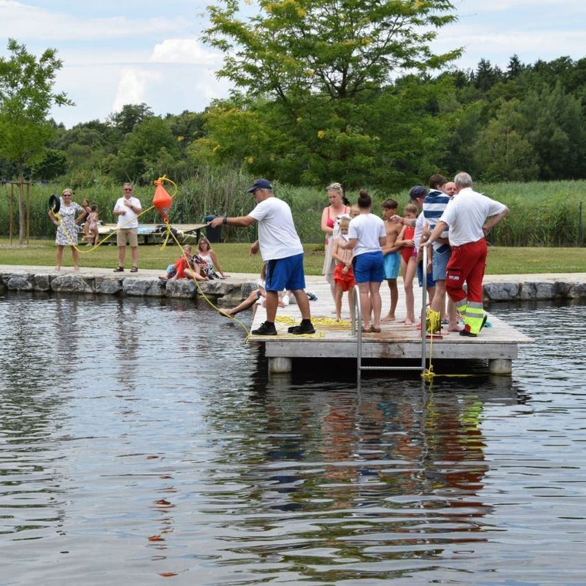 Eine Gruppe von Menschen versammelt sich auf einem Steg am See. Einige tragen Hüte, Sonnenbrillen und Schwimmwesten. Sie beobachten eine Person, die eine Boje hält, möglicherweise im Begriff, eine Wasseraktivität zu beginnen.