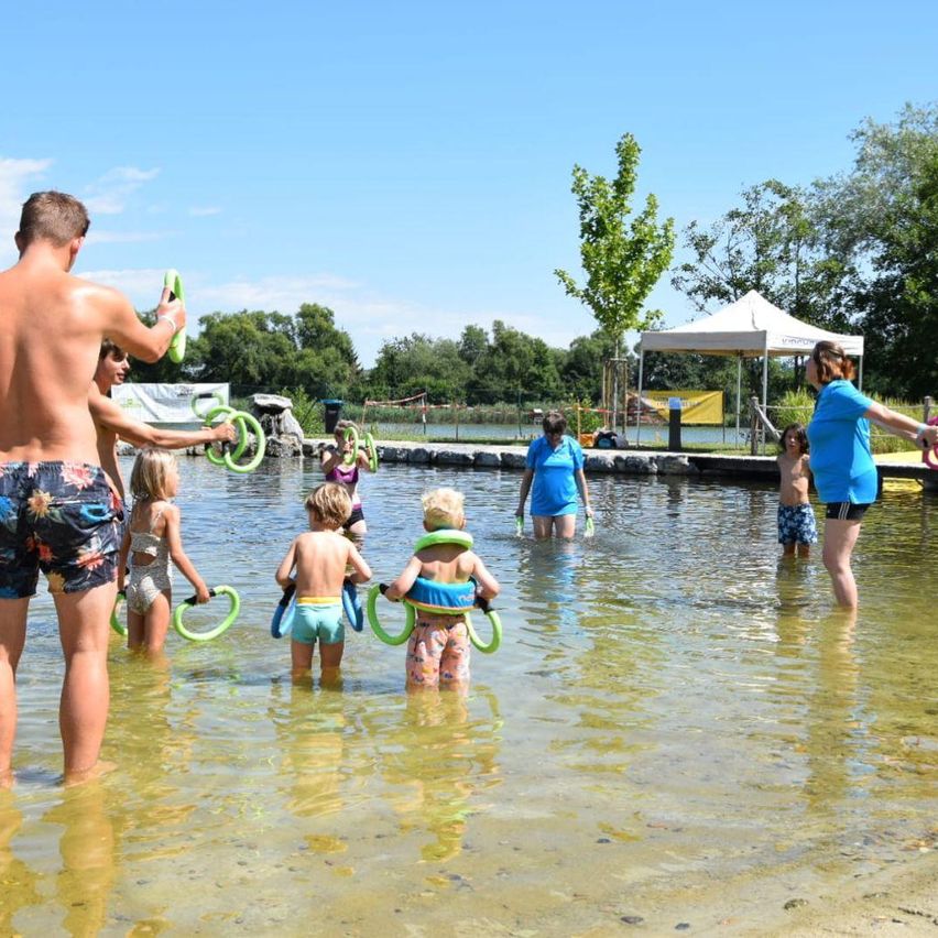 Eine Gruppe von Erwachsenen und Kindern ist in einem See, einige tragen Schwimmringe. Ein Erwachsener hält einen grünen Ring, während eine Frau in der Nähe eines Zeltes steht. Bäume und ein entfernter See sind im Hintergrund zu sehen.