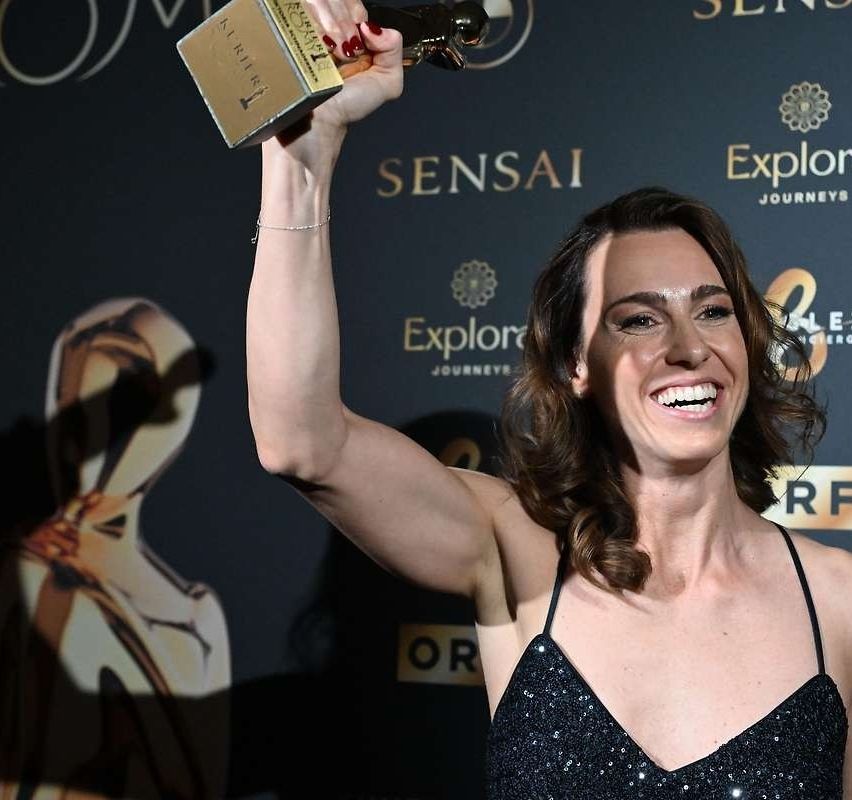 Eine Frau lächelt und hält eine goldene Auszeichnung in der linken Hand. Sie trägt ein schwarzes Kleid und einen Armreif. Sie scheint sich bei einer Preisverleihung zu befinden. Die Wand hinter ihr hat Logos und Text, einschließlich 'Explore Journeys'.