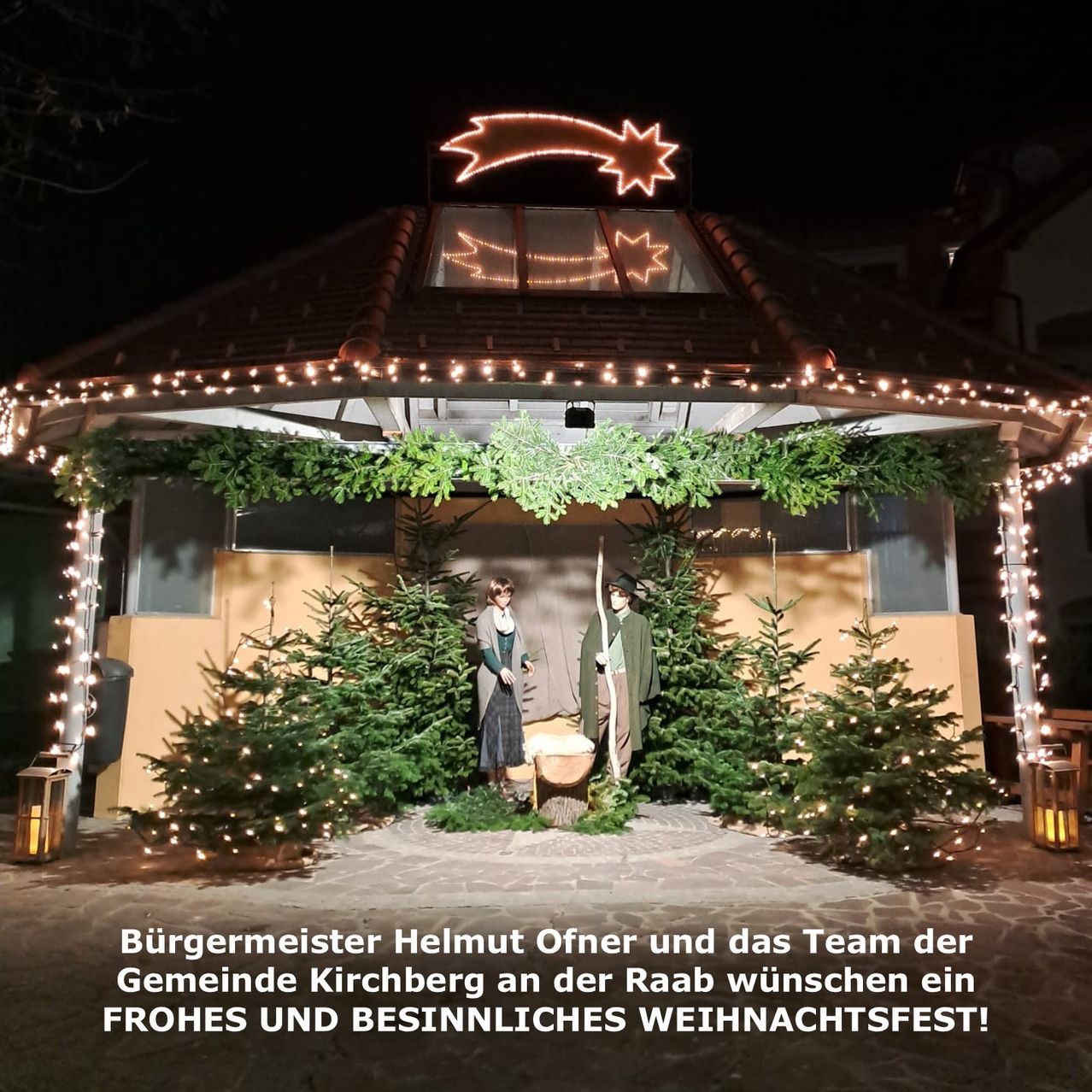 Eine Krippe ist mit Lichtern und Bäumen vor einem Pavillon geschmückt. Zwei Personen stehen in der Mitte. Darunter steht geschrieben: 'Bürgermeister Helmut Ofner und das Team der Gemeinde Kirchberg wünschen Ihnen ein frohes und besinnliches Weihnachtsfest!'