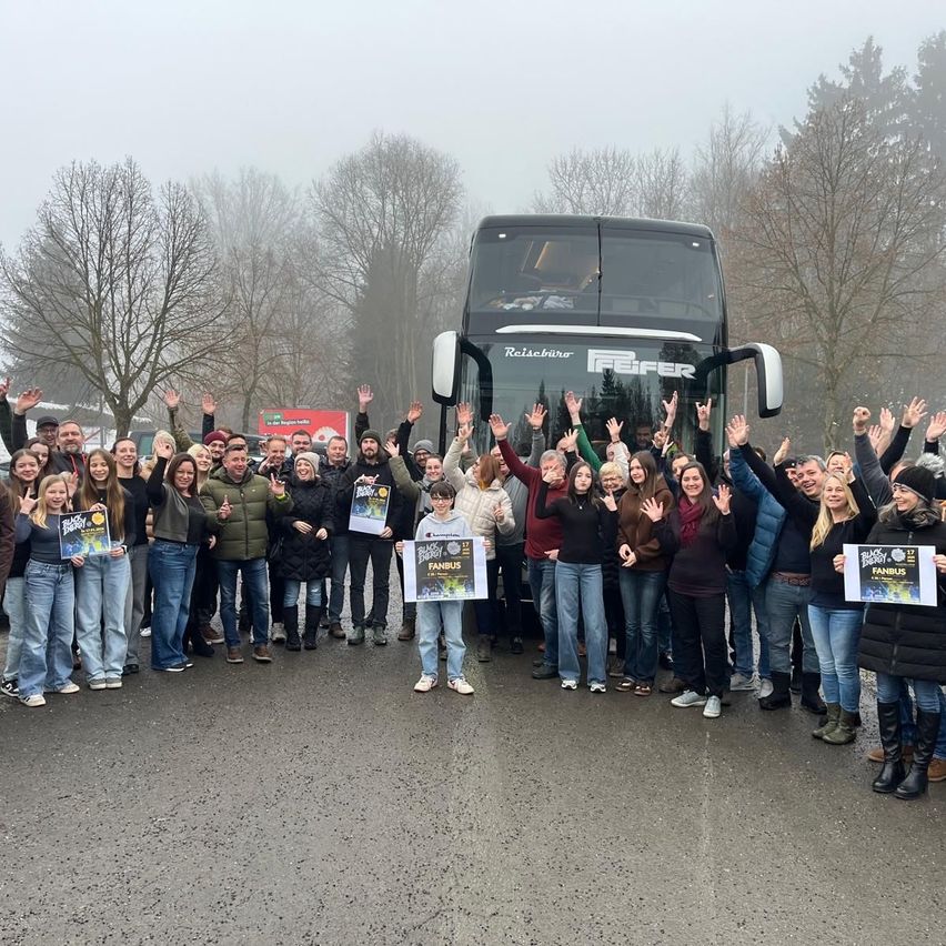 Eine Gruppe von Menschen steht vor einem Doppeldeckerbus, lächelt und hebt die Hände. Sie halten Schilder mit dem Wort Fanbus. Der Bus ist auf einer Straße geparkt, mit Bäumen und einem roten Banner im Hintergrund.