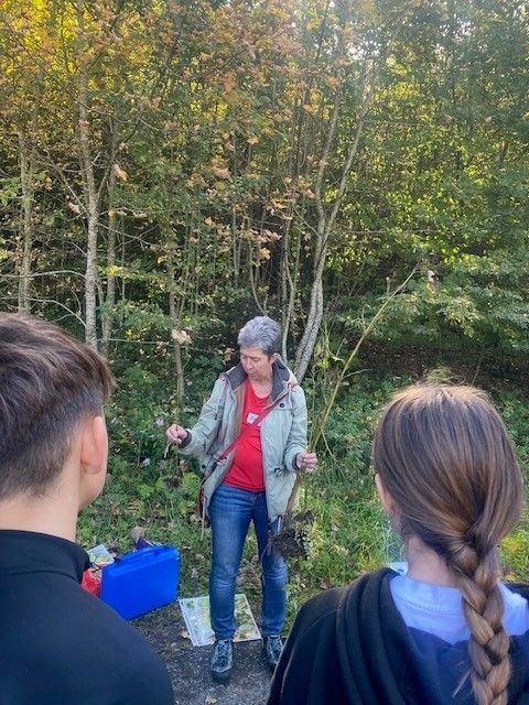 Eine Frau in einem roten Shirt und einer Jacke unterrichtet zwei Schüler in einem Wald und hält einen Stock. Ein blauer Koffer liegt neben ihnen auf dem Boden.
