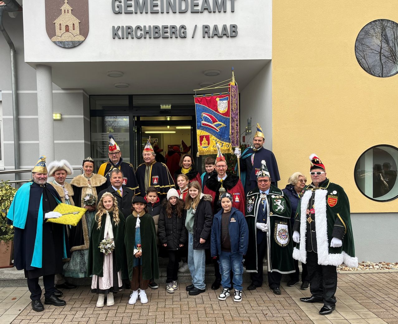 Eine Gruppe von Menschen in Kostümen steht vor dem Gemeindeamt Kirchberg/Raab und posiert für ein Foto. Einige tragen Kronen, und ein Banner mit einem Wappen hängt hinter ihnen.