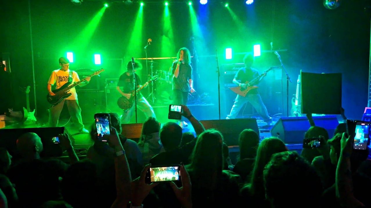 Eine Band tritt auf einer Bühne mit grüner und blauer Beleuchtung auf. Der Leadsänger steht in der Mitte, mit zwei Gitarristen auf beiden Seiten. Das Publikum hält den Moment mit ihren Handys fest.