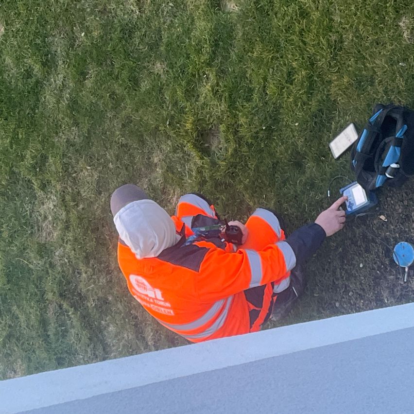 Ein Arbeiter in einer orangefarbenen Sicherheitsjacke sitzt auf dem Gras und hält ein Gerät. Verschiedene Werkzeuge sind um ihn herum verstreut.