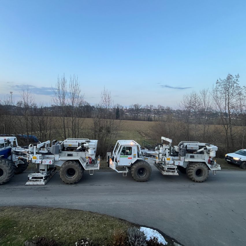 Drei große Maschinenfahrzeuge sind am Straßenrand geparkt, mit einem kleinen Auto dahinter. Bäume umgeben das Gebiet, und ein Feld erstreckt sich im Hintergrund.