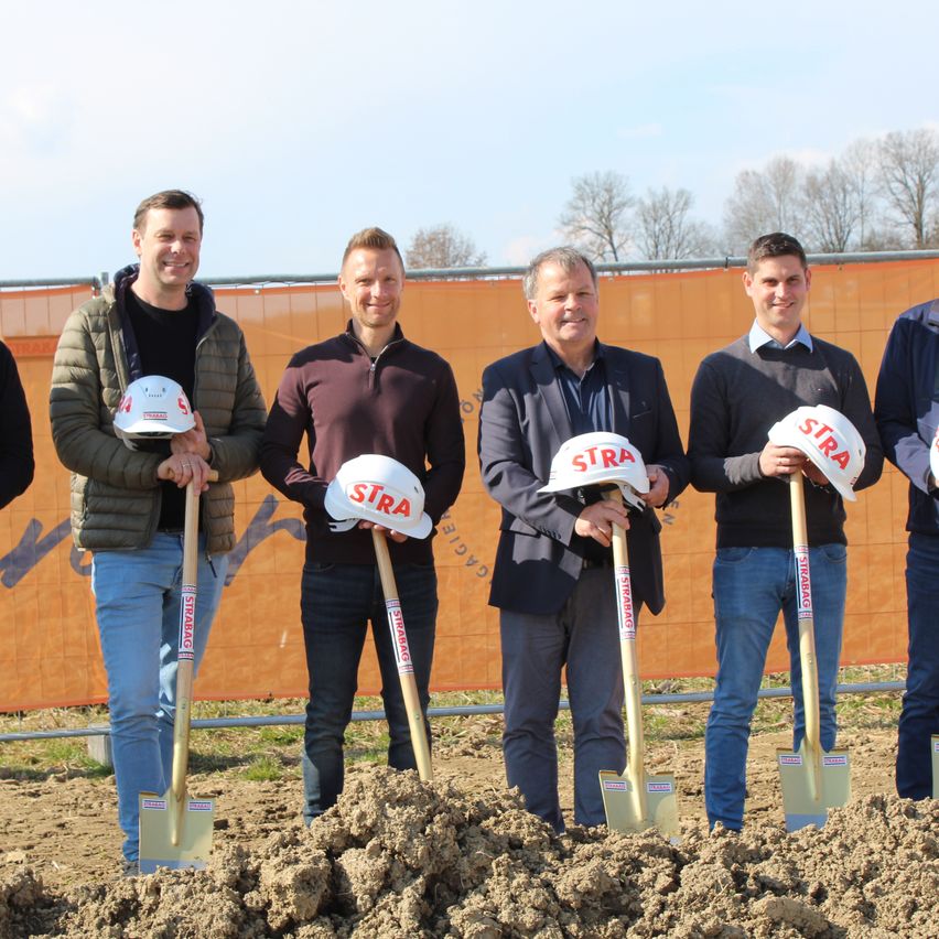 Fünf Männer halten Schaufeln und tragen Helme mit der Aufschrift STRABAG, sie stehen in der Erde vor einem orangefarbenen Banner.