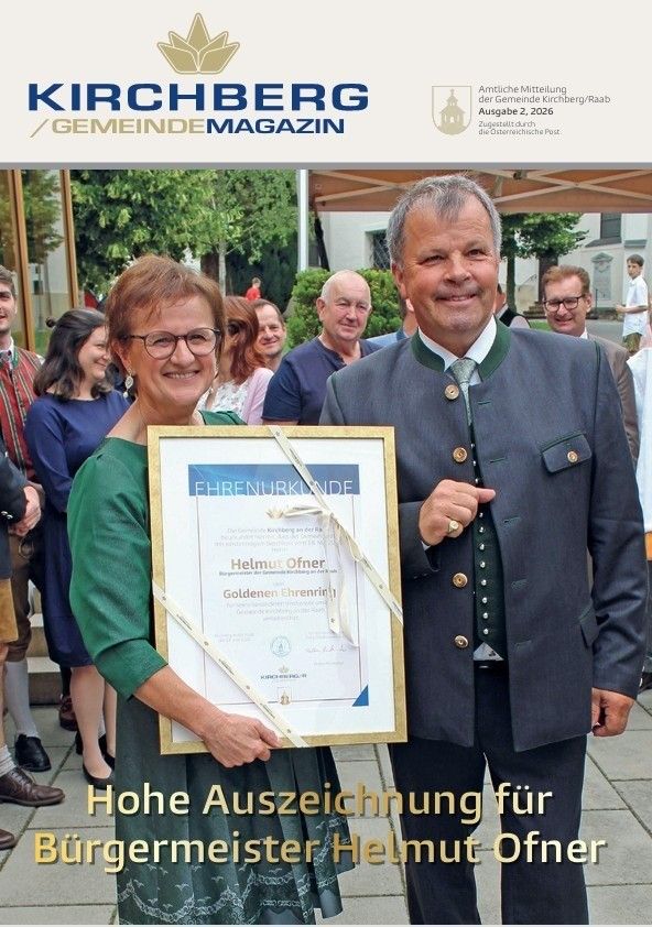 Ein Mann und eine Frau stehen lächelnd nebeneinander und halten eine gerahmte Urkunde. Der Mann trägt ein traditionelles deutsches Outfit und die Frau trägt eine Brille und ein grünes Kleid. Hinter ihnen stehen mehrere Personen, einige mit Brille und formeller Kleidung. Das Bild stammt aus der Kirchberg Gemeinde Zeitschrift vom 2. August 2026.