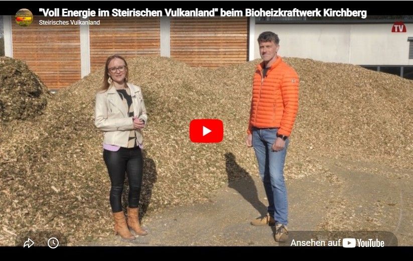 Zwei Personen stehen vor einem Haufen Holzspäne, eine hält ein Mikrofon in der Hand und die andere trägt eine orange Jacke. Der Hintergrund zeigt eine Holzstruktur.