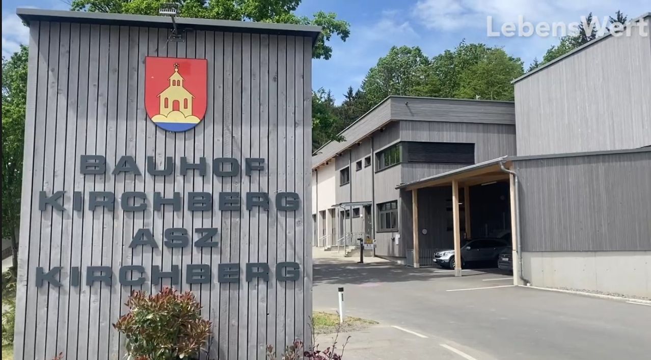 A sign reading 'Uhof chberg aasz chberg' stands on a wooden wall with a logo. Behind it, a building with glass windows and a car parked in front. Trees and a cloudy sky in the background.