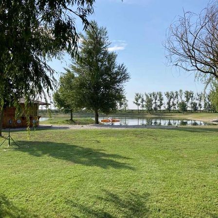 Eine friedliche Außenszene mit einem Teich und einer kleinen Struktur auf der linken Seite. Bäume und Büsche umgeben den Teich, mit einem klaren blauen Himmel darüber.