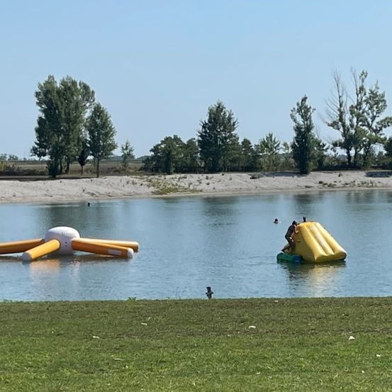 Ein Freiluft-Wasserpark mit aufblasbaren Attraktionen auf einem ruhigen See mit Bäumen und Gras im Hintergrund.