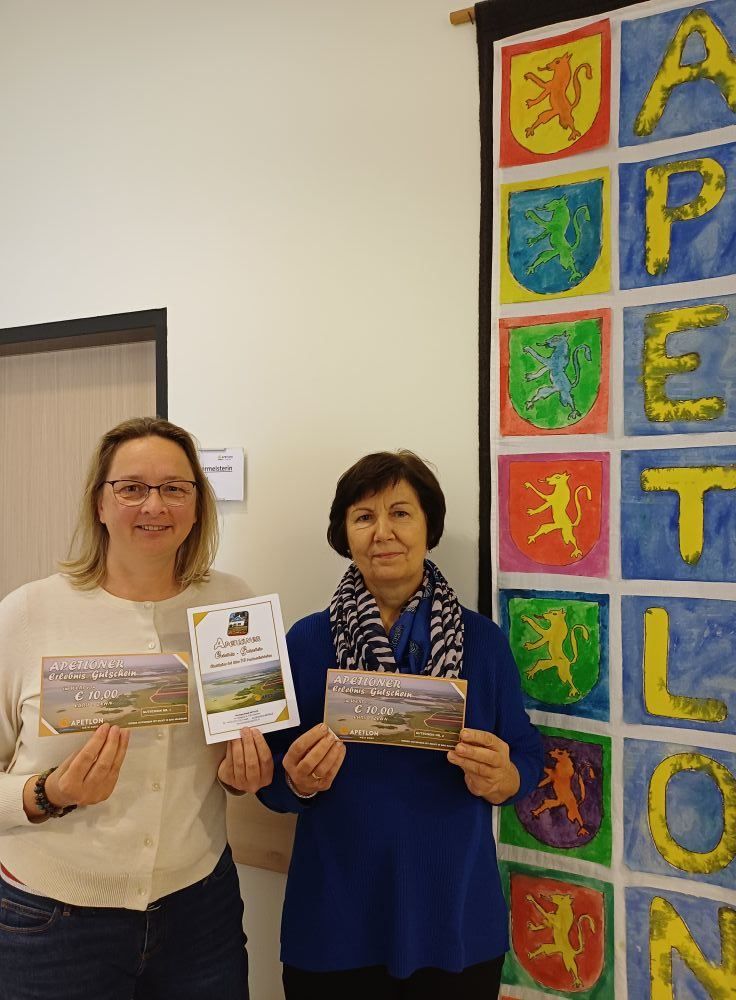 Zwei Frauen stehen vor einer bunten Wand, jede hält eine Karte mit Text und Bildern. Die Karten zeigen eine Landschaft und einen Preis von 10,00 €. Die Wand hat mehrere Quadrate mit unterschiedlich gefärbten Löwen und dem Wort PETLO.