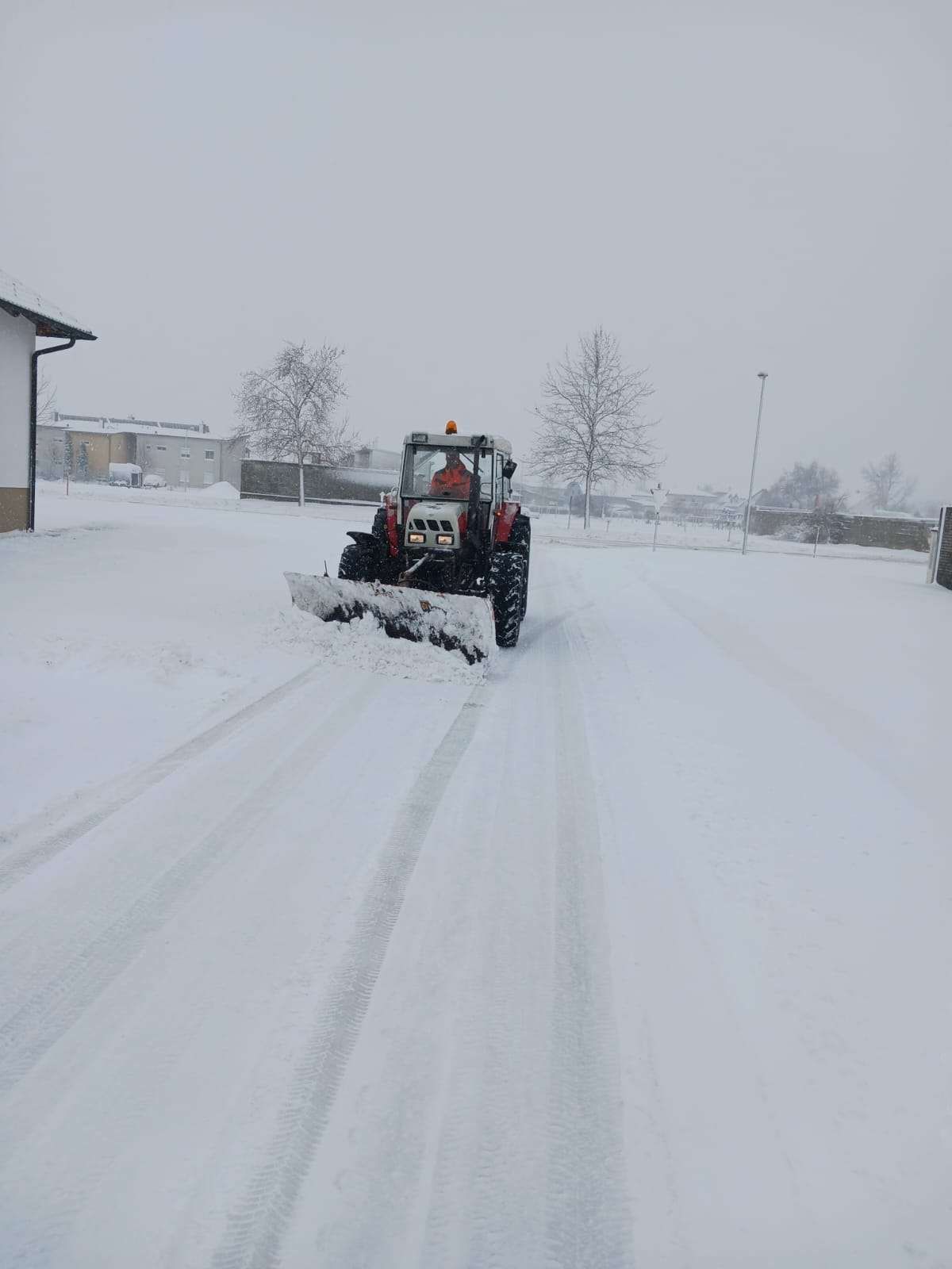 Ein Traktor mit Schneepflugaufsatz räumt Schnee von einer Straße. Im Hintergrund sind Bäume und Gebäude zu sehen.